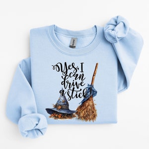 May include: A light blue crewneck sweatshirt featuring a graphic print of a dark blue witch hat, a brown broomstick, and the black cursive text "Yes, I can drive a stick!" The design includes small orange autumn leaves around the base of the hat.