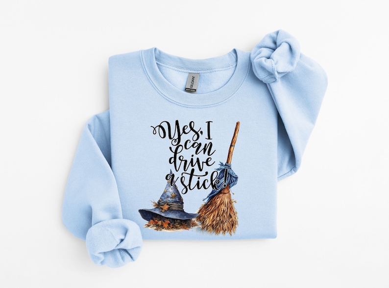 May include: A light blue crewneck sweatshirt featuring a graphic print of a dark blue witch hat, a brown broomstick, and the black cursive text "Yes, I can drive a stick!" The design includes small orange autumn leaves around the base of the hat.