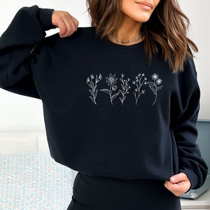 May include: A black long-sleeve sweatshirt featuring a white line art graphic of five distinct wildflowers arranged in a horizontal row across the center of the chest. The garment has a crew neckline and ribbed cuffs on the long sleeves.