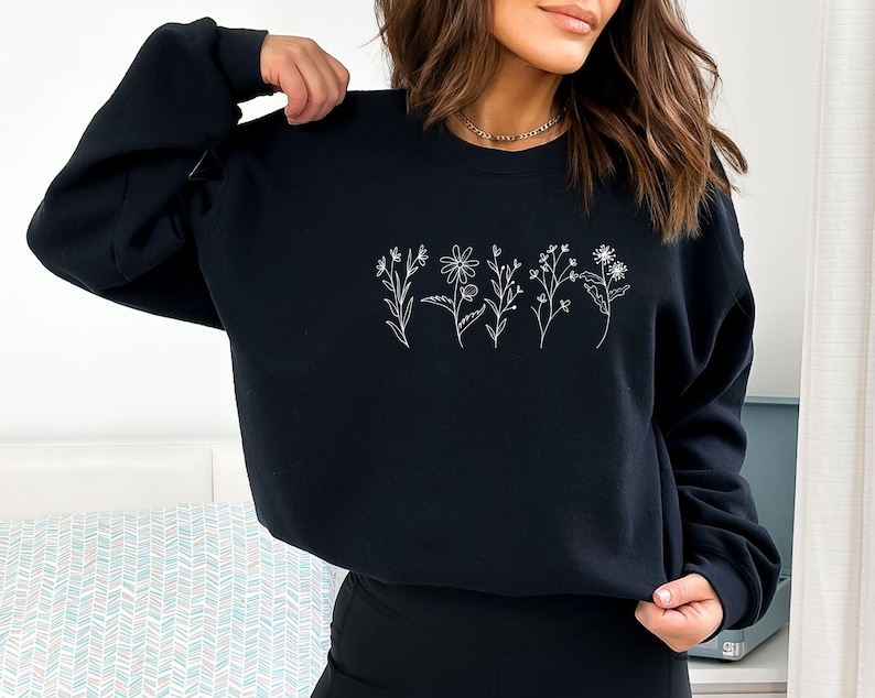 May include: A black long-sleeve sweatshirt featuring a white line art graphic of five distinct wildflowers arranged in a horizontal row across the center of the chest. The garment has a crew neckline and ribbed cuffs on the long sleeves.