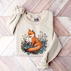May include: A beige crewneck sweatshirt featuring a graphic illustration of an orange fox sitting among various blue, yellow, and pink flowers and green leaves. The Gildan brand tag is visible at the inner neckline of the long-sleeved garment.