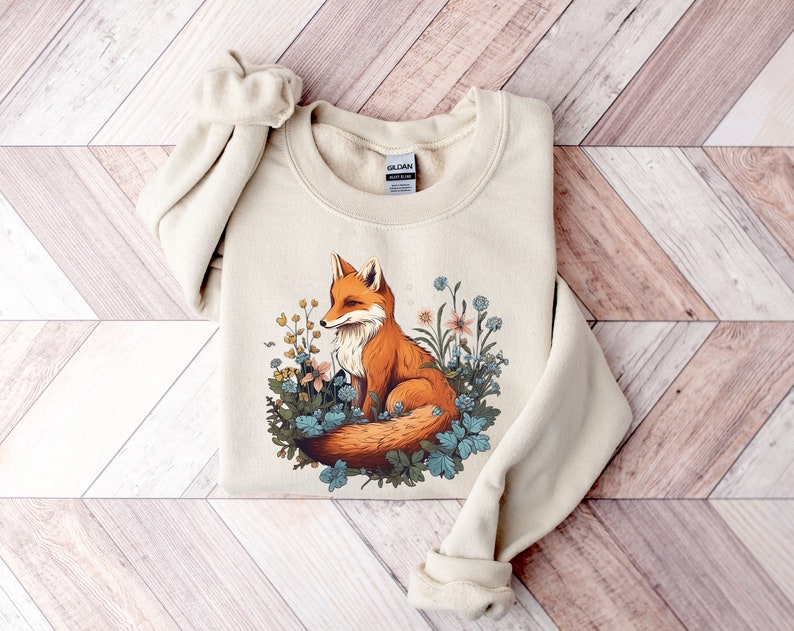 May include: A beige crewneck sweatshirt featuring a graphic illustration of an orange fox sitting among various blue, yellow, and pink flowers and green leaves. The Gildan brand tag is visible at the inner neckline of the long-sleeved garment.