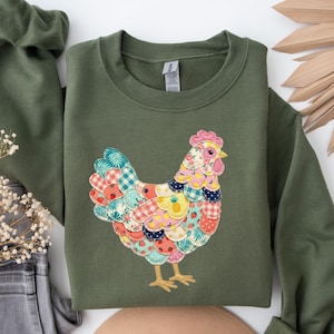 May include: Olive green sweatshirt featuring a colorful patchwork chicken design. The chicken has a pink comb, yellow feet, and a body made of various patterned fabrics. The sweatshirt has long sleeves and a crew neck.