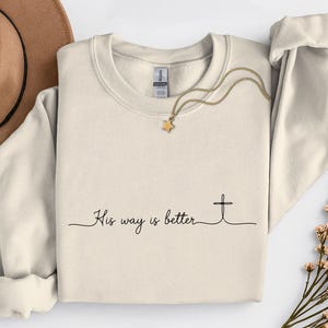 May include: Cream-colored sweatshirt with the text "His way is better" and a cross graphic. A gold necklace with a star pendant and a brown hat are also visible. The sweatshirt has long sleeves and a crew neck.