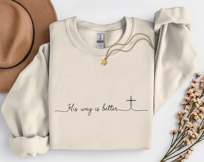 May include: Cream-colored sweatshirt with the text "His way is better" and a cross graphic. A gold necklace with a star pendant and a brown hat are also visible. The sweatshirt has long sleeves and a crew neck.