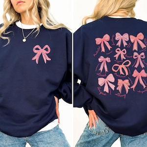May include: A navy blue crewneck sweatshirt featuring a small pink bow graphic on the front left chest. The back displays a collection of various pink bows accompanied by the words Protected, Loved, Unique, Strong, Precious, Brave, and Valued.