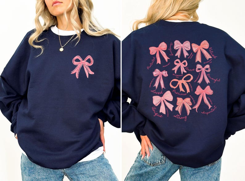May include: A navy blue crewneck sweatshirt featuring a small pink bow graphic on the front left chest. The back displays a collection of various pink bows accompanied by the words Protected, Loved, Unique, Strong, Precious, Brave, and Valued.