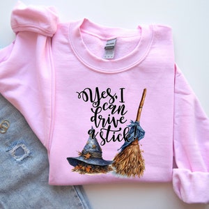 May include: A light pink long-sleeve sweatshirt featuring a graphic of a blue witch hat resting on autumn leaves next to a straw broom. The black text reads, Yes, I can drive a stick! The garment has a crew neckline and ribbed cuffs.