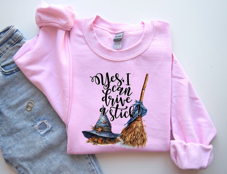 May include: A light pink long-sleeve sweatshirt featuring a graphic of a blue witch hat resting on autumn leaves next to a straw broom. The black text reads, Yes, I can drive a stick! The garment has a crew neckline and ribbed cuffs.