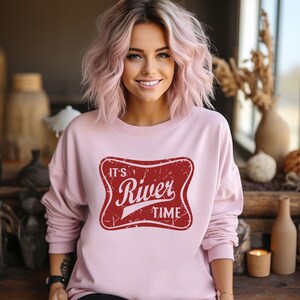 May include: A light pink long-sleeve sweatshirt featuring a central red rectangular graphic with rounded corners. The graphic has a distressed texture and contains the white text "IT'S River TIME" in a stylized, bold, and script font combination.