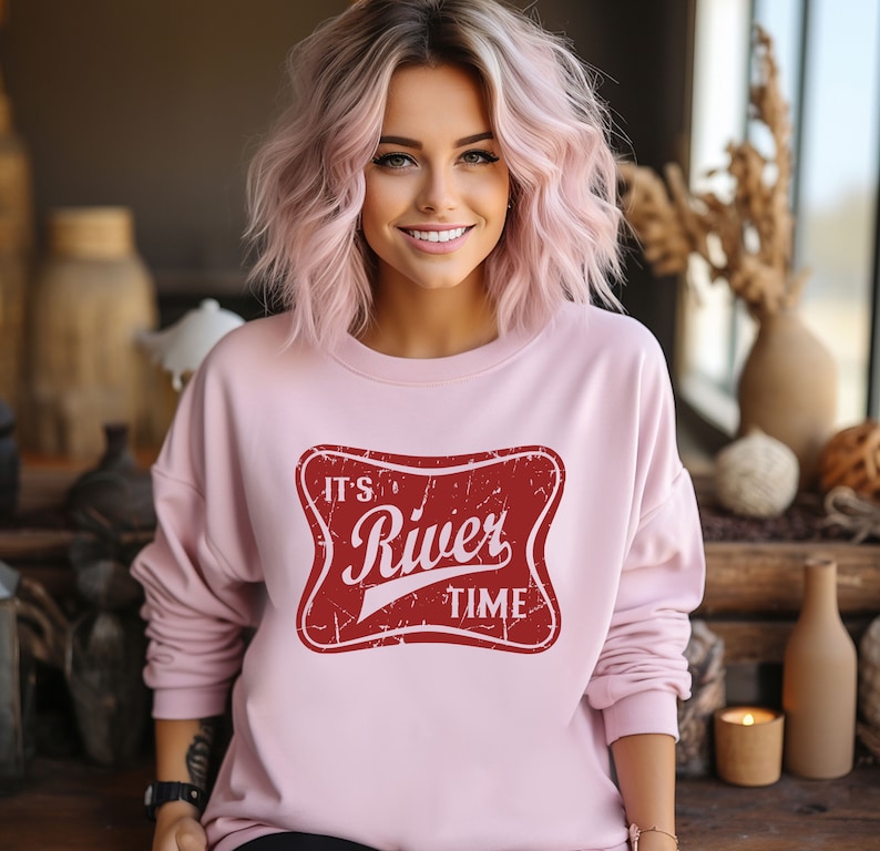 May include: A light pink long-sleeve sweatshirt featuring a central red rectangular graphic with rounded corners. The graphic has a distressed texture and contains the white text "IT'S River TIME" in a stylized, bold, and script font combination.