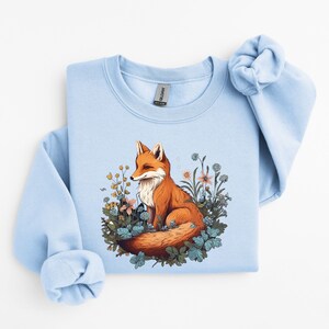 May include: A light blue crewneck sweatshirt featuring a graphic illustration of an orange fox sitting amidst a cluster of colorful wildflowers and green foliage. The design is centered on the chest of the garment, which has long sleeves.
