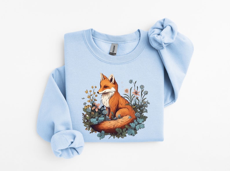 May include: A light blue crewneck sweatshirt featuring a graphic illustration of an orange fox sitting amidst a cluster of colorful wildflowers and green foliage. The design is centered on the chest of the garment, which has long sleeves.