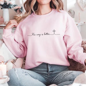 May include: A light pink sweatshirt with the text "His way is better" and a cross design in black. The sweatshirt is a crew neck style and appears to be made of a soft material.