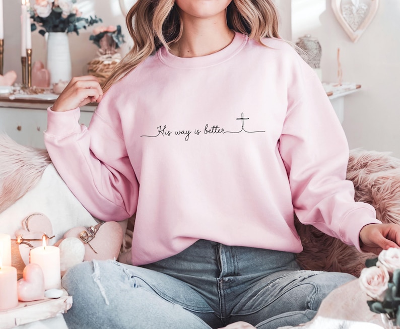 May include: A light pink sweatshirt with the text "His way is better" and a cross design in black. The sweatshirt is a crew neck style and appears to be made of a soft material.