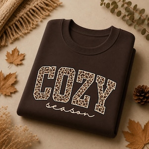 May include: A dark brown crewneck sweatshirt featuring the word COZY in large block letters with a leopard print pattern. Below the word COZY, the word season is written in a white cursive script font. The garment has a ribbed collar.