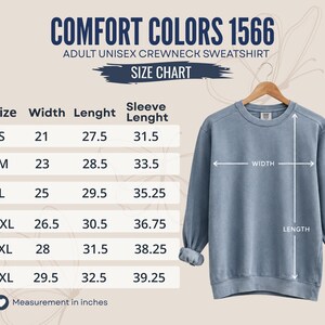 K&ouml;nnte beinhalten: Ein blaues Sweatshirt mit Rundhalsausschnitt auf einem Holzkleiderb&uuml;gel neben einer Gr&ouml;&szlig;entabelle mit dem Titel Comfort Colors 1566 Adult Unisex Crewneck Sweatshirt. Die Tabelle listet Gr&ouml;&szlig;en von S bis 3XL mit Ma&szlig;en in Zoll f&uuml;r Breite und L&auml;nge.