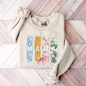 Pu&ograve; includere: Una felpa beige con un design floreale e la parola "MAMA" in lettere bianche. Il design floreale &egrave; in tonalit&agrave; di blu, giallo, rosa e verde.
