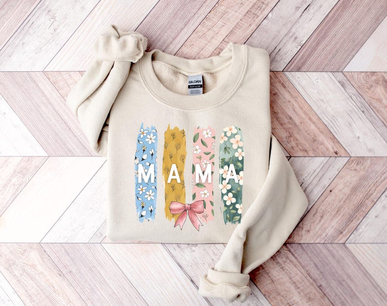 Pu&ograve; includere: Una felpa beige con un design floreale e la parola "MAMA" in lettere bianche. Il design floreale &egrave; in tonalit&agrave; di blu, giallo, rosa e verde.