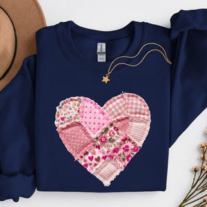 Pode incluir: Camisola azul marinho com um design de coração em patchwork. O coração é composto por vários tecidos rosa e estampados florais. Um colar de ouro com um pendente de estrela está drapeado sobre a camisola. Um chapéu castanho é visível no fundo.