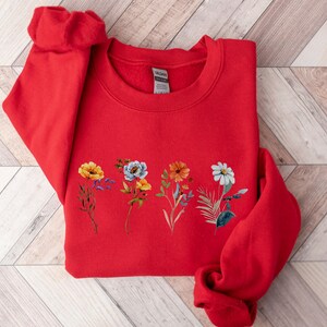 May include: Red sweatshirt with a floral design featuring yellow, orange, white, and blue flowers.