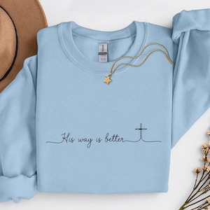 May include: A light blue crewneck sweatshirt with the text "His way is better" and a cross graphic. A gold necklace with a star pendant is draped across the sweatshirt. A brown hat and floral sprigs are in the background.