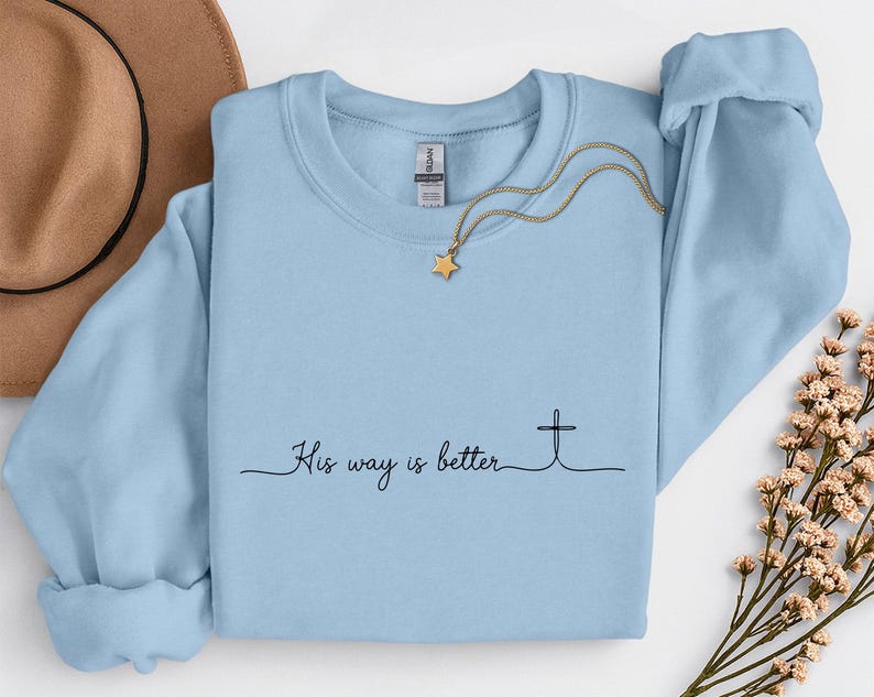 May include: A light blue crewneck sweatshirt with the text "His way is better" and a cross graphic. A gold necklace with a star pendant is draped across the sweatshirt. A brown hat and floral sprigs are in the background.