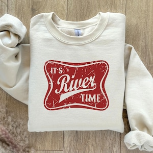Puede incluir: Una sudadera beige con un gráfico rojo desgastado que dice "It's River Time".