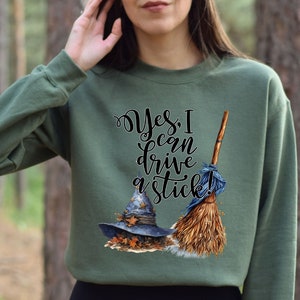 May include: An olive green crewneck sweatshirt featuring a graphic print of a blue witch hat resting on autumn leaves next to a broomstick. The text "Yes, I can drive a stick!" is printed in black cursive and serif lettering above the illustrations.