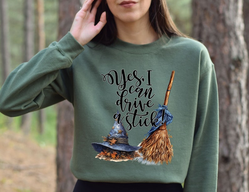 May include: An olive green crewneck sweatshirt featuring a graphic print of a blue witch hat resting on autumn leaves next to a broomstick. The text "Yes, I can drive a stick!" is printed in black cursive and serif lettering above the illustrations.