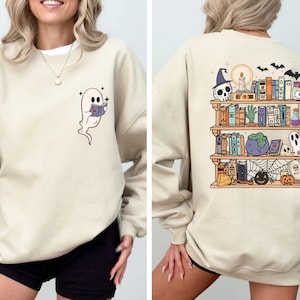 May include: Beige sweatshirt with a cartoon ghost reading a book on the front. The back features a bookshelf with Halloween-themed items like skulls, bats, and pumpkins. A cozy and festive design for the spooky season.