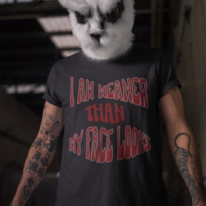 May include: A person wearing a black t-shirt with the text "I am meaner than my face looks" in red. The person is wearing a white rabbit mask with black ears. The person has tattoos on their arms.