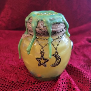 May include: A small glass jar with a green wax lid and a string wrapped around the top. The jar contains a yellow liquid and has two charms hanging from it: a star and a crescent moon.
