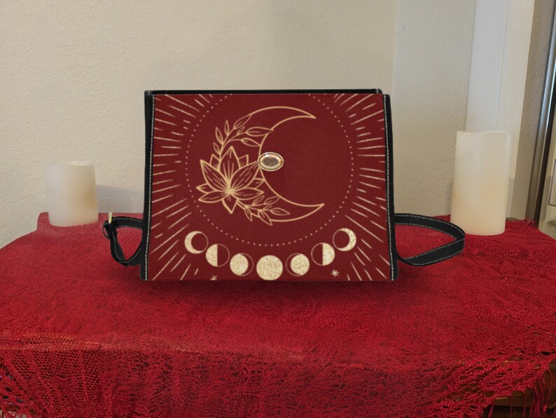 Pode incluir: Uma bolsa tote vermelha com um design de lua dourada e flores. A bolsa tem uma al&ccedil;a preta e um design de fases lunares douradas.
