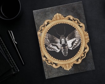 May include: A notebook featuring a detailed watercolor illustration of a Death's-head Hawkmoth within an ornate gold frame. The moth is depicted in shades of gray and white against a black background. The notebook has a dark gray cover.