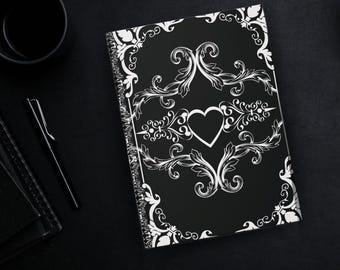 Puede incluir: Cuaderno negro con un corazón blanco y un diseño floral. El cuaderno está encuadernado en espiral y presenta un borde decorativo. Un bolígrafo y una taza negra son visibles en el fondo.