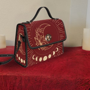 Pode incluir: Uma bolsa de m&atilde;o de couro sint&eacute;tico vermelho com uma al&ccedil;a preta e um design de lua e flores douradas. A bolsa tem uma aba superior com um fecho dourado.