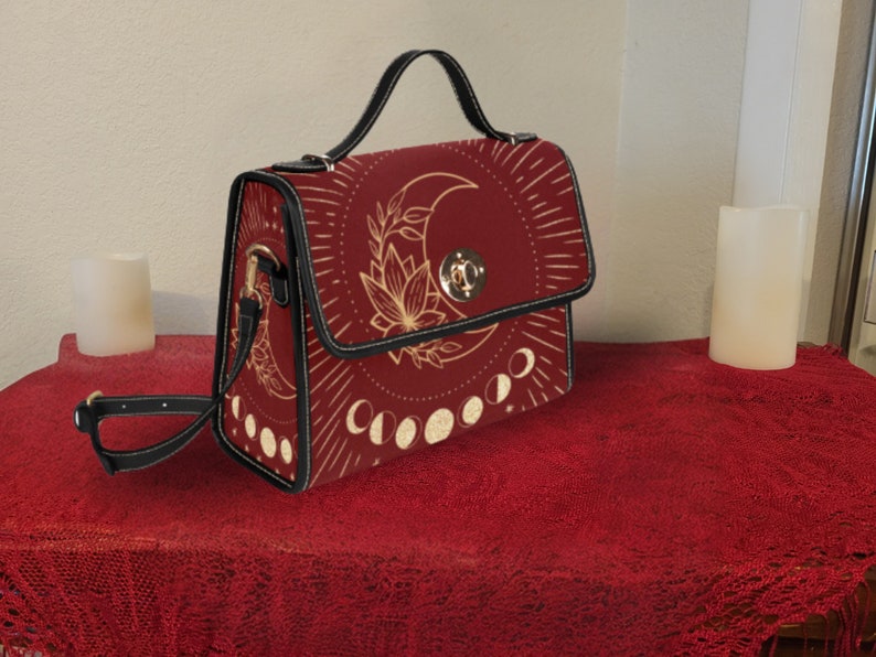 Pode incluir: Uma bolsa de m&atilde;o de couro sint&eacute;tico vermelho com uma al&ccedil;a preta e um design de lua e flores douradas. A bolsa tem uma aba superior com um fecho dourado.