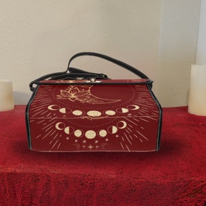 Pode incluir: Uma bolsa de couro vermelha com uma al&ccedil;a preta e um design de lua e estrelas douradas. A bolsa tem um fecho de aba e uma al&ccedil;a superior.