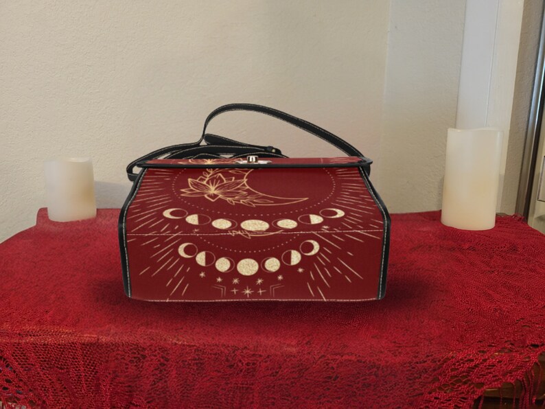 Pode incluir: Uma bolsa de couro vermelha com uma al&ccedil;a preta e um design de lua e estrelas douradas. A bolsa tem um fecho de aba e uma al&ccedil;a superior.