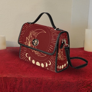 Pode incluir: Uma bolsa de ombro de couro sint&eacute;tico vermelho com uma al&ccedil;a preta e um design de lua e estrelas douradas. A bolsa tem uma al&ccedil;a superior e um fecho de aba.