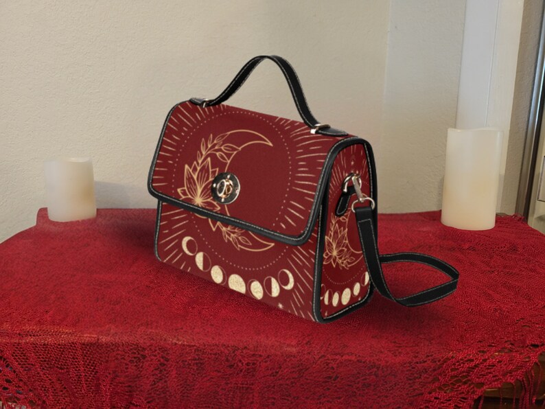 Pode incluir: Uma bolsa de ombro de couro sint&eacute;tico vermelho com uma al&ccedil;a preta e um design de lua e estrelas douradas. A bolsa tem uma al&ccedil;a superior e um fecho de aba.