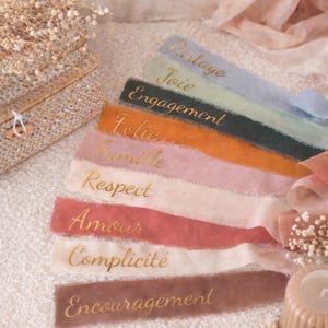Peut inclure: Rubans colorés avec des inscriptions dorées, dont les mots "Engagement" et "Amour". Les rubans sont disposés en diagonale sur un fond doux et texturé. La palette de couleurs comprend des nuances de bleu, vert, orange, rose et marron.