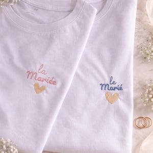 Peut inclure: Deux t-shirts blancs avec du texte brodé et une icône de cœur. L'un affiche le texte rose "la Mariée" au-dessus d'un cœur doré, tandis que l'autre affiche le texte bleu "le Marié" au-dessus d'un cœur doré, centrés sur la poitrine.