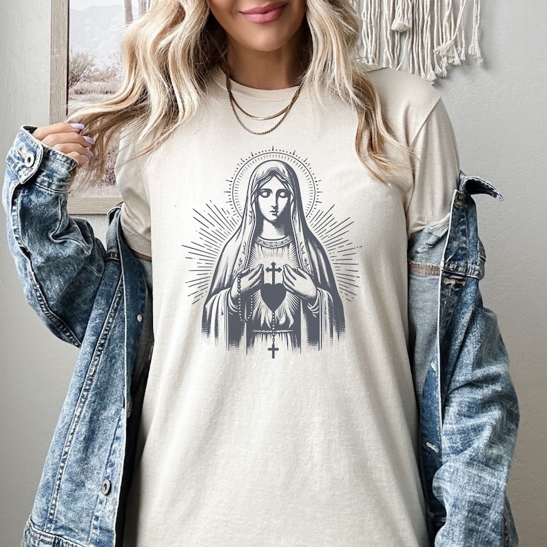 May include: A white t-shirt featuring a graphic of the Virgin Mary holding a heart with a cross on it. The graphic is in shades of grey and black.