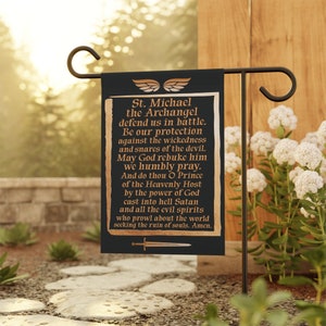 May include: Black garden flag with gold text that reads "St. Michael the Archangel defend us in battle." The flag has a gold border and a sword graphic. The flag is hanging on a black metal stand.