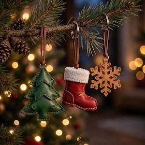 May include: Three hanging ornaments attached to a pine branch: a green leather Christmas tree, a red leather boot with a white faux fur cuff, and a tan leather snowflake, each suspended by a brown leather strap and a metal ring.