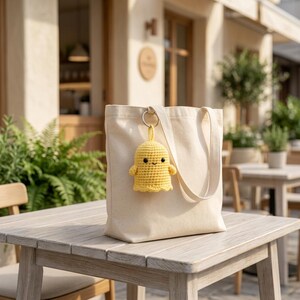 May include: A plain off-white canvas tote bag with two shoulder straps. Attached to one of the straps by a silver metal ring is a small, handmade yellow crochet ghost figure featuring a simple stitched face with two black eyes and a smile.