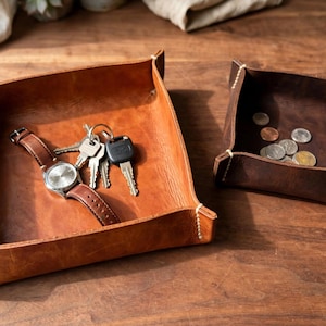 May include: Two square leather valet trays with stitched corners. The larger tan tray holds a silver wristwatch with a brown leather strap and a set of metal keys. The smaller dark brown tray contains several assorted silver and copper coins.