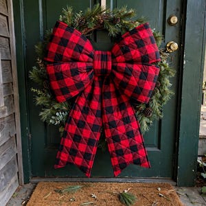 May include: A circular evergreen wreath featuring a large, quilted bow with a red and black buffalo plaid pattern. The wreath is constructed from pine branches and small pinecones, centered with the prominent fabric bow.